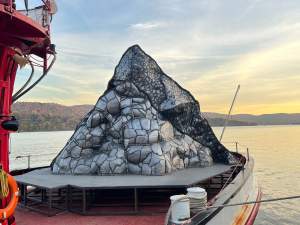 Photographed on the JJ Harvey fireboat in the Hudson River during its trajectory from Storm King Art Center to Troy, NY under a yellow- and blue tinted sky, a pyramidal sculpture sits on a metal-footed plinth in the bow of a boat. The sculpture is made up of silvery gray steel bulges outlined with black. Some sections fit together like irregular cobblestones and other areas, especially at the top, are made up of smaller sections outlined in black.