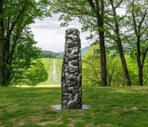 A welded steel monolith stands in a grassy area at Storm King Art Center in front of trees that open onto a distant view of fields and hills under a cloudy sky. The shiny but textured surface of the metal form is separated into sections, which are encased in black lines, creating the impression of stones stacked as in a fieldstone wall.