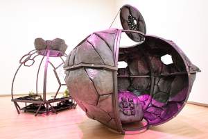 A pod-like welded steel free-standing sculpture in the shape of a space capsule is partially open on one side and is lit with fuchsia-pink light. Sitting just behind the pod, a second structure made up of metal beams create a ball-shaped cage in which plants grow, a space garden, carrying plants and a carved stone with a formula for the distribution of seeds.