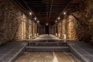 A long room with rough stone walls and a flat, wood-beamed ceiling is mostly filled with platforms made of steel grids. Metal beams create loose cages around six tanks, three to each side. In each tank is a sculpture of a person’s face and cloak-like hair draping over the shoulders. The heads all face inward and are made of a white material.