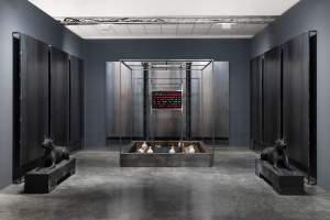 In a room with a gray concrete floor and charcoal-gray walls, trios of metal panels stand against each of the three walls in our view. Steel-gray sculptures of Doberman dogs flank the space to either side of a metal cage, which nearly fills the height of the room. At least eight white amphora or vases are set in the base, and a screen above them has green or red text against a black background.