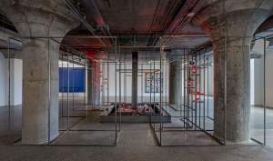 Metal cages encase four squat, upward-flaring concrete columns in a low-ceilinged space. The metal structures surround a fifth, which has a tray-like square base set with ceramic vases around a central metal beam.
