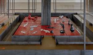 Closer view of the central tray-shaped platform with ceramic and clay vessels, cast human hands, and statuettes that seem to melt in slow motion. An angular trough surrounds the central platform and a second trough outside of that is filled with brown water.
