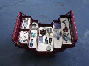 Twenty-one objects are arranged nestled into or on top of five silver, reflective levels of a red cantilevered toolbox. The box opens to create a chunky V, and objects include bottles and tubes, including an Oxi Clean pen, a brush, flossers, and a pair of metal clawed bird’s feet.