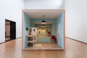 View into a box constructed in a white-walled gallery. Inside the box are light blue walls, a wooden countertop table, white refrigerator with photos, a box of Special K cereal, a fold-down table or ironing board, and a wooden chair set under a black hanging lamp. The rear wall opens onto five ever-smaller rooms with exactly the same furnishings.