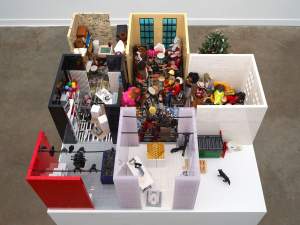 View down into a doll-scaled structure of seven rooms and two open spaces, which together make a grid of three-by-three cubes. One room holds a urinal laid down on a table and other materials, and another is set up as an office.  At least 19 dolls line an L-shaped space created by three cubes. The dolls have different colors of hair and skin, and each is elaborately dressed.
