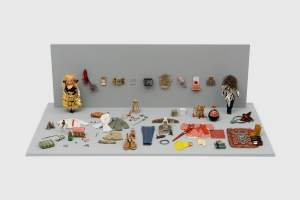 Two dove-gray panels are set up to make a back wall and floor of a diorama scattered with doll-sized items of clothing, shoes, and accessories. Two dolls stand to either side near the back wall, bookending a hung display of miniature materials and sculptural pieces. One doll has light-toned skin and blond hair, and wears a skirt made up of gold purses. The others face is hidden by a tangled cloud of brown hair.
