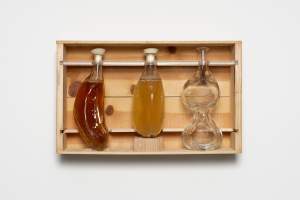 Three clear glass, organically shaped vessels are displayed in a wooden case. The leftmost is roughly half-moon shaped and is filled with amber brown liquid. The central vessel is a lopsided oval filled with pale yellow liquid, and the rightmost has two bulbs, one over the other like connected blobs in a lava lamp. The third vessel is empty, and the other two have white stoppers.
