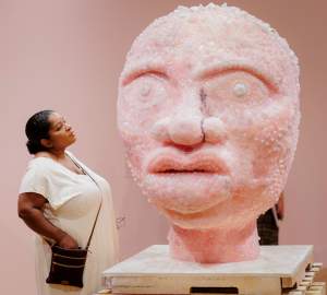 A round, pink head made up of pieces of rose quartz and a short neck sit on a wood palette base in a gallery. The sculpture has wide eyes with flower-shaped pupils, a rounded nose with one nostril split away from the rest, and full, protruding lips. The base of the sculpture comes up to hip-height of a person standing next it, and the viewer’s face comes to the level of the sculpture’s nose.