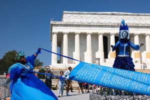 The artist wears a voluminous blue gown and flowered headpiece as she pulls two ribbons attached to a long piece of sky-blue cloth. Words are written in all-capital, gold letters on the cloth, which is pulled over a structure like a parade float holding an abstracted mannequin with Marian Anderson’s face printed in blue-toned black and white.