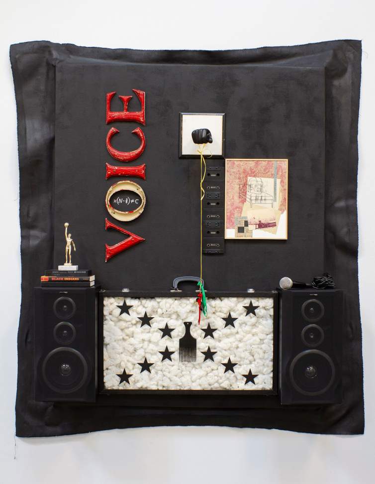 Speak, so you may speak again is a  mixed-media wall assemblage featuring the word "VOICE" in red, speakers, a microphone, a framed sketch, and various audio equipment mounted on a black backdrop. The lower section has a rectangular field of cotton texture, overlaid with an x of stars and an afro pick at the center.