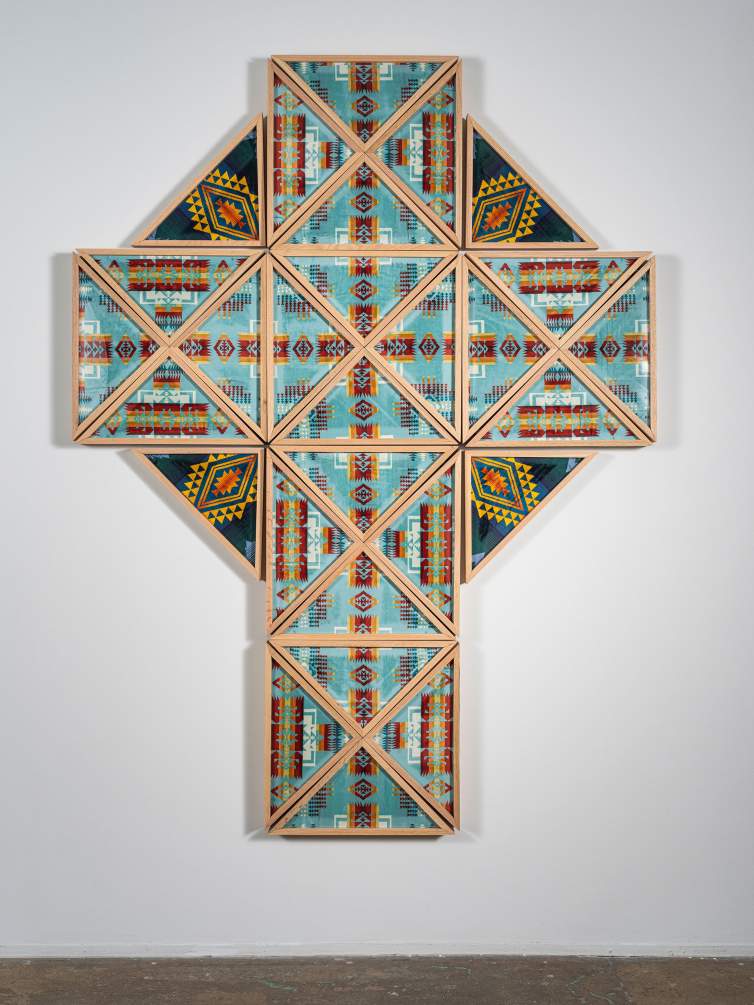 Here Comes the Son is a wall-mounted art piece in the shape of a cross composed of triangular ceremonial flag frames holding Pendleton geometric patterned beach towels.