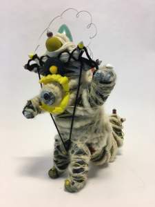 Claude is a small figure-like sculpture wrapped with yarn. Yellow vampire costume teeth outline a snout, and small arms are extended up. A crown is formed with yarn and objects, and a scalloped wire haloes its head.