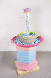 Fountain of Your Youth is a fountain formed of a plastic tube with a rotating spiral pushing white and blue plastic pony beads up to the top, where they spill out into the stacked bowls below, a fluted concrete bowl lined on the inside with lime green, and a wide pink colander below that, both filled with accumulated pony beads. The fountain sits on a column of pink, blue and orange pool noodles coiled to look like a column, wrapped with transparent mesh.