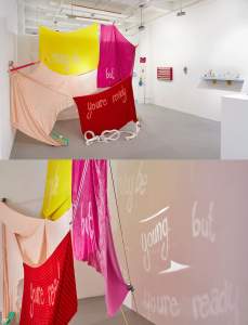 i may be young, but youre ready is a sculptural installation in a white-walled gallery, with 5 rectangles of velvet in light pink, yellow, fuschia and red, suspended and clipped together and to an oversized pink pencil puncturing the wall. The surface of the velvet has been burned out to read “ I may be young but youre ready.” A light is cast through the fabric, and the words also read on the wall behind it. On the floor is a white coiled conveyor belt, and a tan and green geometric object that holds down a corner of the light pink velvet.