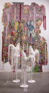 they wondered what to do … is a brocade and sequined installation with two figures, floral patterns and dripping with beads, accompanied by white roosters on pedestals in front draped with white beads.