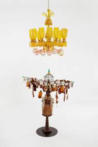 Lady of commerce is a sculpture with an amber glass chandelier, festooned with white eggs laced with red. Below, a monk-like figure is on a stand, with a wire structure and bird cage above him from which brown bottles hang with other objects.