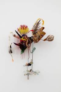 Addictions to leaf and nut aroused… is an abstract assemblage hanging on a white wall, formed with feathers, small bottles, fabric, glass beads, thread, and dried gourds.