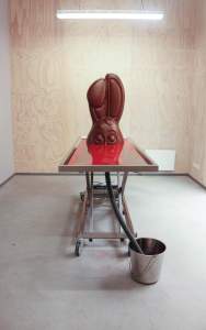Bliss Point (Saint Gabriel's Morgue) is an installation of a stainless steel rolling embalming table with the top half of a larger-than-life chocolate bunny face standing upright in a pool of bright red fluid. A flexible drain pipe extends from the table to a metal bucket.