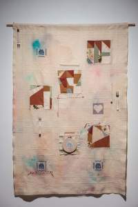 Homebound is a beige grid-textured textile hung on the wall, with 4 interstitial fragmented quilt squares, embroidered geometric houses, and 2 knives and forks wrapped with embroidery floss.