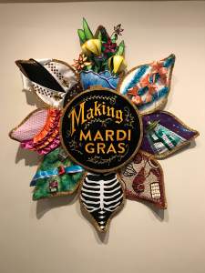 A spangled embroidery hoop at the center reads “Making Mardi Gras” in embroidered decorative letter, and 8 large petals extend from the hoop, each with an iteration of decorative costume elements, including silk ruffles, papier-mache flowers, sequins, seed art, and a painted ribcage.