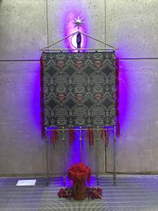 Portable Monument to Harriet Tubman — True American Hero is a sculpture centered on a grey and white square painting with a repeating moiré pattern held in decorative fence-like stand. At the top, is a triangle finial with an open hand in the center and a star extending from the top. Red cords fringe the sides and are piled on a stand in front, along with a pile of dried roses.