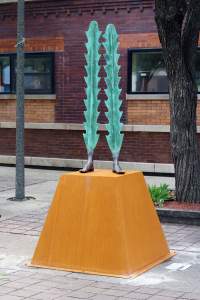 Caminantes is a sculpture in a public plaza. A pair of feet stand on a weathered golden-brown steel base. From the metal feet, two green cactus-like shapes extend upward.