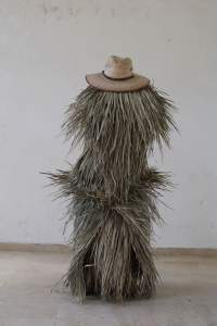 Kajne: Untitled I is an image of a performance where a figure is entirely covered with palm leaves, topped with a brimmed hat.