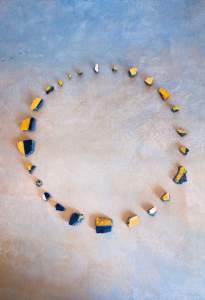 Aurora is a photography of rock fragments of various shape and size, painted with gold and blue arranged in a circle on a stone like beige and blue mottled surface.