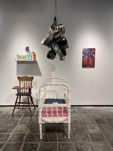 Soy de una raza pura is an installation of various objects in a museum space. A cluster of metal cookware hangs above a white crib with stars-and-stripes sheets. Behind it a high chair sits below a shelf with various containers, and a painting of two girls hangs to the side.