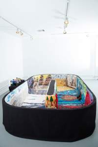 Snake Tube Adventure Racing is a low oblong racetrack, the outside covered in black fabric, and the interior lined with various quilted landscapes. 3 small toy-like wheeled vehicles are lined up next to the track.