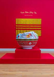Chén is a sculpture on a red stepped plinth, of an oversize replication of plastic “Longevity” chinese-style bowls, with a band of red decoration, and rim of yellow and black interlocking decoration. In the bowl are labeled bags of various rice, and behind on a red wall is a yellow floral textile banner with 3 stripes of red, and the text on the wall reads “Tram Nam Song Tho Dinh Viet Anh Dai”.