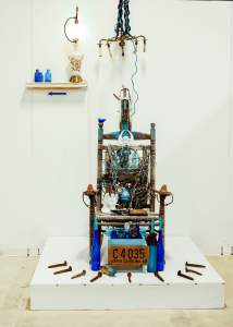 An artistic installation featuring a chair adorned with various wires, bottles, a license plate, and other eclectic items, set against a plain wall.