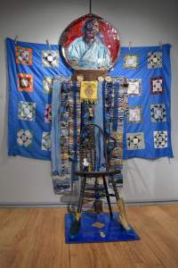 Art installation with a chair, a colorful quilt backdrop, a circular portrait on top, and various items like fabric strips, a glass bottle, and a shoe are attached.