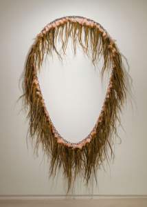 RAT KING is an oval art piece made of long, brown, hair-like strands intertwined with chunky flesh-like silicone, mounted on a wall.