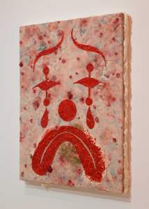 Cry Now is a mixed media piece of a sad face in red on a textured, multicolored background, made out of pink mottled silicone with red face features.