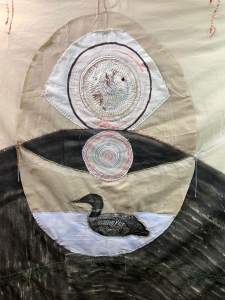 Loon Protector is a textile artwork featuring stitched concentric circles above an embroidered loon swimming on water, with neutral and dark fabric creating an abstract background.