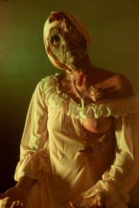 On the Way Back shows a person in a pale, off-shoulder gown and head covering, a drooping green face with prosthetic wounds and fingers emerging from their face, chest, and shoulder, sits under dramatic yellow-green lighting with one breast exposed.