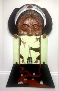 Que Brillaba Sobre El Muro is a mixed-media artwork depicting a sculptural large human face with braids and an open mouth at the top, from which a painting extends with small human figures and a red river flowing toward miniature houses on a dark surface.