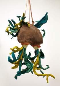 A textile sculpture with green and yellow plant-like forms extends from a corduroy butt hanging by ropes.
