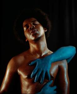 A shirtless person with closed eyes is touched by a blue-painted hand against a dark background.