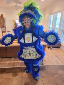 Denise Smith smiles brightly in an elaborate royal blue feathered costume, with large central panels of intricate beadwork, and round panels on her hands. She is a Black woman, and wears her hair in two long black braids under a beaded and feathered headdress.