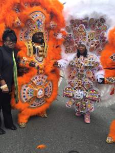 Two people stand on the street in elaborate feathered and beaded costumes. On left, a Black man wears an orange suit, and Black woman on the right wears a white feathered suit with colorful patterned beaded covering the whole front and the elaborate headdress.