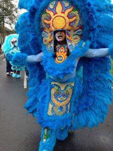 In this photograph titled The Snake, Gang Flag ZeeBo wears a vivid blue suit made up of a head piece with a gold and red sun, a gold and blue skirt, and an oval aura of feathers around the person’s whole body, from high overhead to shin. Gang Flag ZeeBo wears textured blue pants, also perhaps feathers, and a light blue shirt under the suit. He walks with arms wide and mouth wide open.
