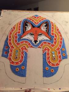 Tiny beads and crystals sewn to canvas create a fox’s face surrounded by bands of sky blue, coral pink, deep yellow, and rows of pearls. Smaller pearls also make tiny rosette-like shapes in the coral-colored band.