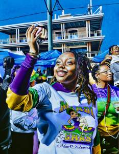 A person with brown skin and long black braided hair holds up a coconut and smiles widely in this painting titled MY FIRST COCONUT. The person wears hoop earrings, a nose ring, and a shirt with Betty Boop on text reading, “Mardi Gras New Orleans.” Other adults and children fill the space between the person and an angular building on the other side of the crowd.