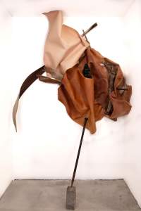 Junto, te aplico is a sculptural installation, here set in a square alcove. A swath of tawny brown leather ripples and flares like a flame against the back and one side wall. A thick tree branch, a long dried palm frond, and other organic material is nestled in the folds. The piece is pinned to the back wall by the back end of a long iron stake. The point of the stake rests on a concrete block on the floor in the center of the alcove.