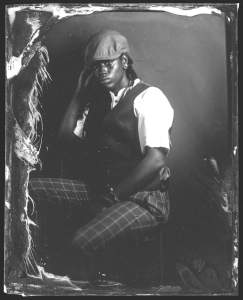 This black and white photograph titled Young Jean in the Studio shows a person sitting with their body angled to our left. They rest one hand between their thighs and the other by their face, which turns to us. The person wears a flat cap, a dark vest over a short-sleeved white shirt, and dark pants with a pinstripe grid. Ripples appear to line the edges of the photo.