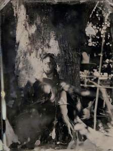 Jean, Loren, and the Oak in the 7th Ward is made up of two overlapping black and white photographs. In one, a young person sits leaning against a tree. Dappled light speckles the person and the brimmed hat they hold. Overlaid to the right, a person shown in a larger scale wears the hat and is partially cut off by the tree.
