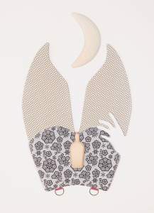 This shaped leather piece, titled Nite Lite, is made up of two pieces. Above, a quilted, ivory-white crescent moon hangs between the long, rabbit-like ears or legs of the lower piece. The ears are covered with a fishnet stocking pattern, and the bottom is patterned with black on gray to create the impression of lace. The shape of a hand is cut out to the lower right, and two hooks hang on the bottom edge. A small, round-shouldered cosmetic bottle is lined up with the opening between the ears, and a pink-colored nub presses out from the end.